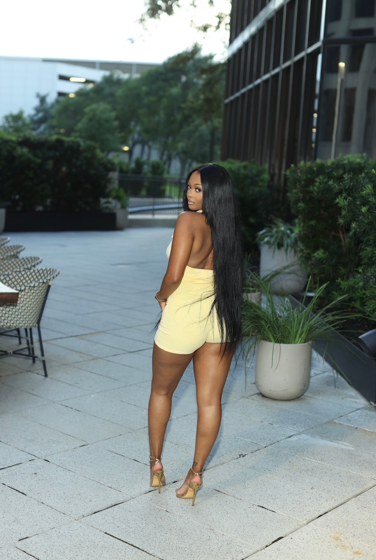 Woman in a yellow outfit standing outdoors on a patio with plants and tables in the background.